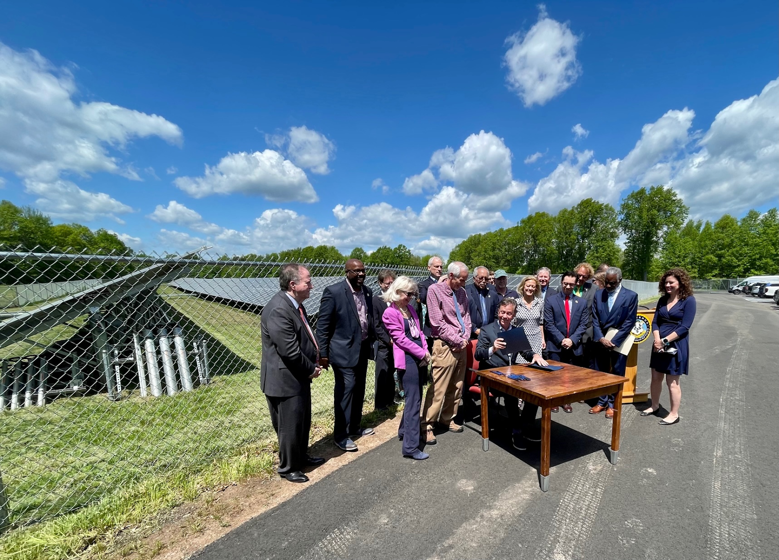 Ned Lamont, signing an executive order outdoors on a sunny day surrounded by happy colleagues 